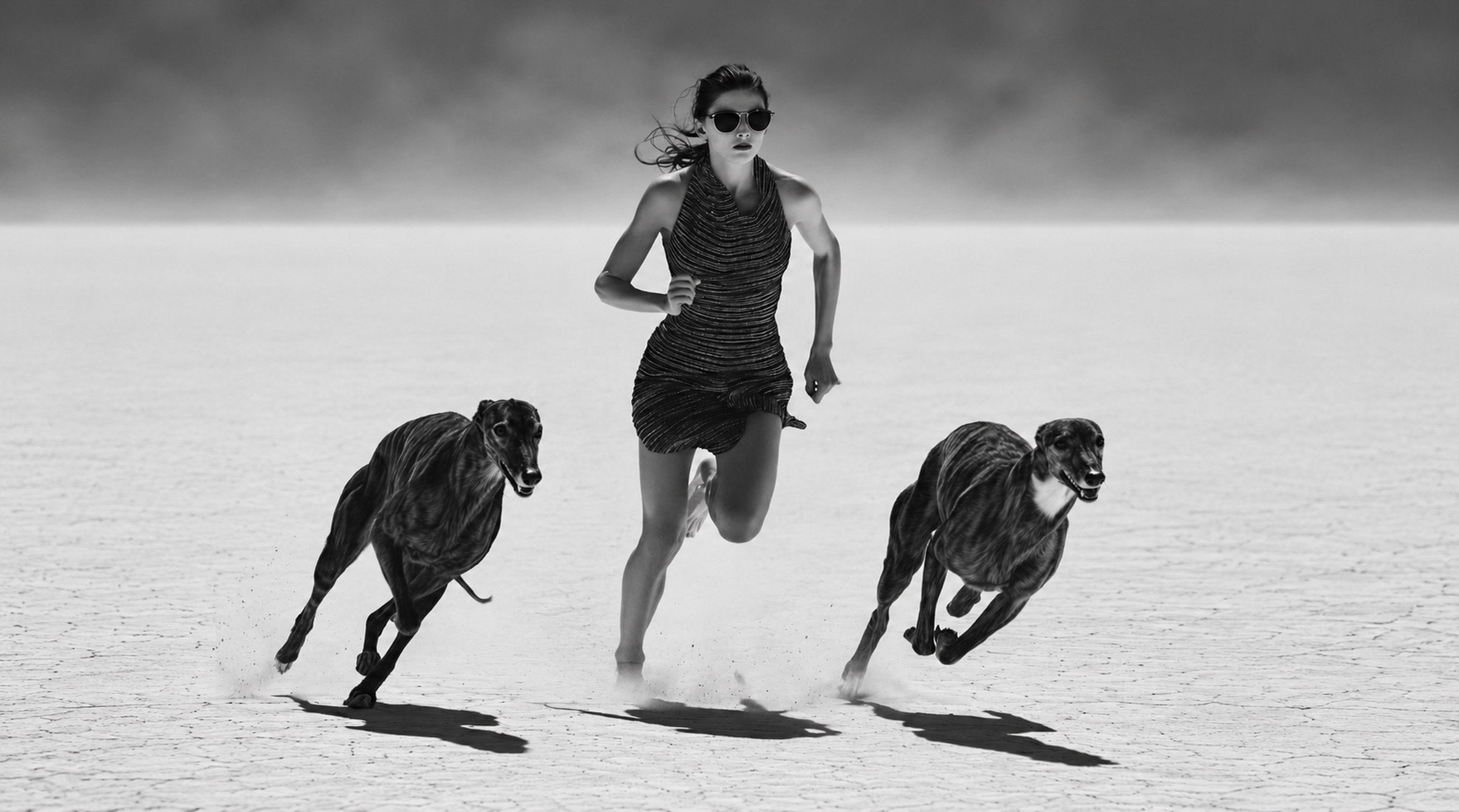 Black and white fashion campaign photograph: a model sprinting barefoot across a cracked dry lake bed, flanked by two greyhounds at full stride